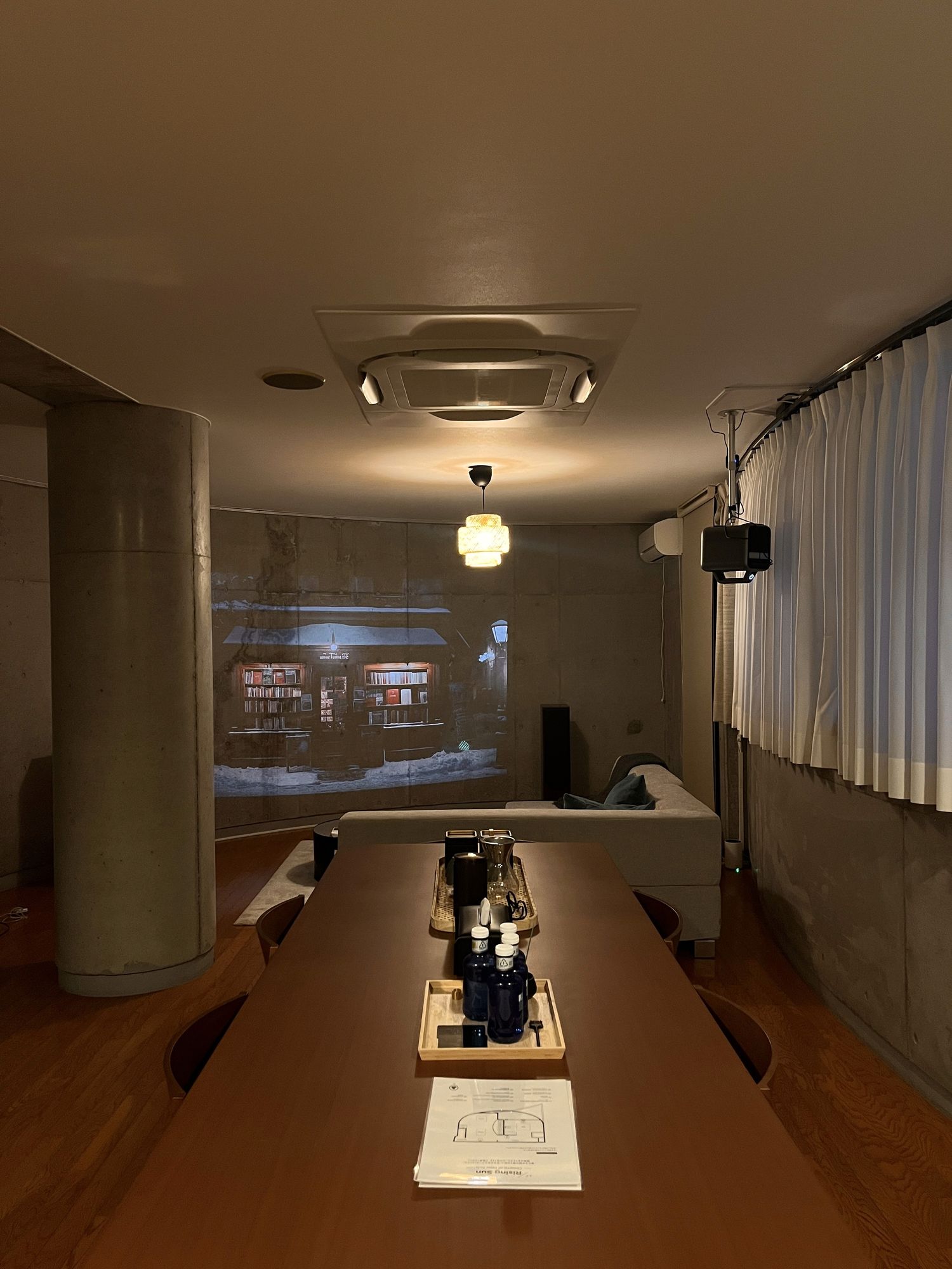 Long timber table beneath a concrete column, night view beyond the window